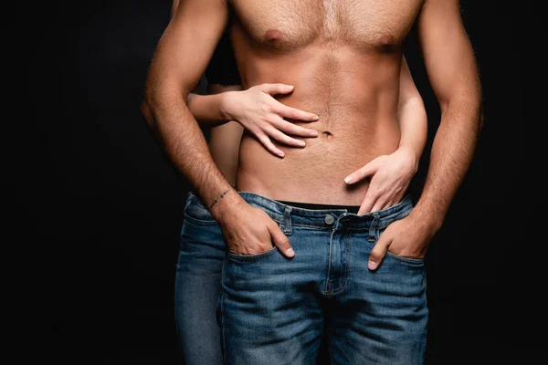 Visão parcial da mulher sedutora abraçando o homem sem camisa com tronco muscular isolado no preto — Fotografia de Stock