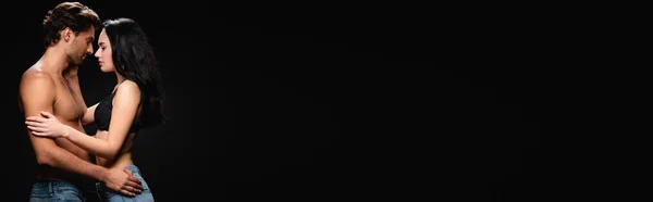 Vista lateral de homem e mulher sem camisa em sutiã abraçando face a face isolado em preto, banner — Fotografia de Stock