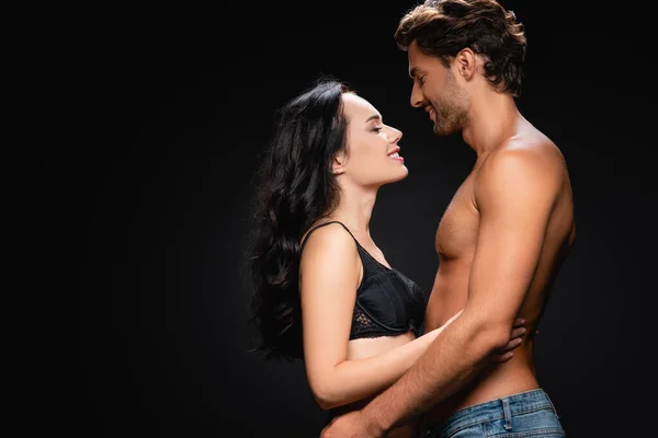 Vista lateral de la feliz pareja joven abrazándose y mirándose aislados en negro - foto de stock