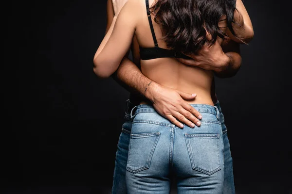 Вид ззаду жінки в джинсах і бюстгальтер біля чоловіка, що обіймає її ізольовано на чорному — стокове фото