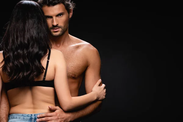 Joven hombre sin camisa mirando a la cámara mientras abraza sexy morena mujer aislado en negro - foto de stock