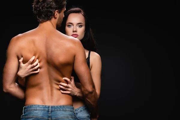 Mujer seductora mirando a la cámara mientras rasca la espalda de un hombre sin camisa aislado en negro - foto de stock
