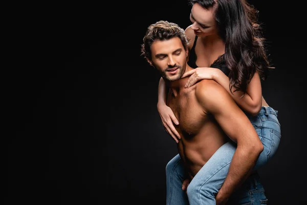 Smiling shirtless man piggybacking sexy woman in bra and jeans isolated on black — Stock Photo