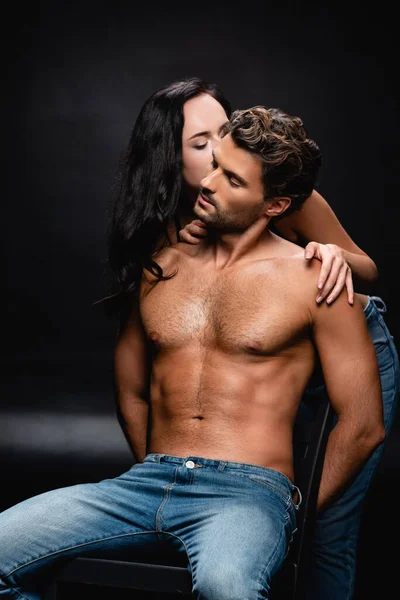 Sensuelle brunette femme câlins et baisers jeune homme musclé assis sur chaise sur noir — Photo de stock