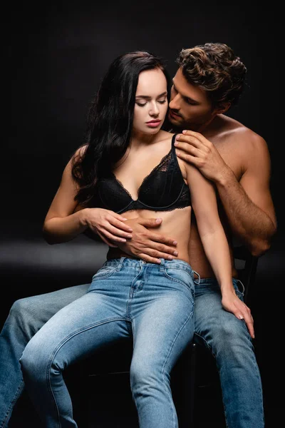 Femme séduisante en jeans et soutien-gorge assis sur une chaise avec un homme torse nu passionné la serrant dans ses bras sur noir — Photo de stock