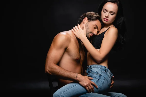 Passionné jeune couple en jeans étreignant les yeux fermés sur chaise sur noir — Photo de stock