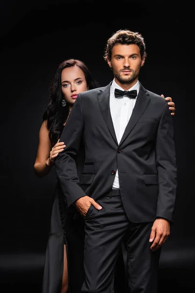 Young confident man with hand in pocket near sensual brunette woman on black — Stock Photo