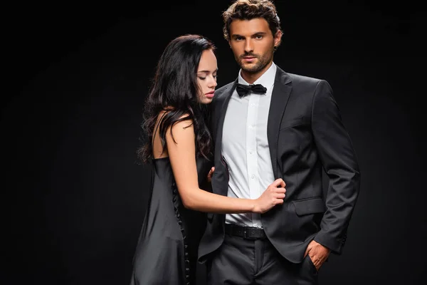 Confident man with hand in pocket looking at camera near seductive woman in satin dress isolated on black — Stock Photo