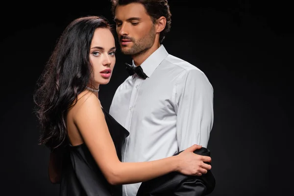 Sexy brünette Frau schaut in die Kamera, während sich eleganter Mann isoliert auf schwarz auszieht — Stockfoto