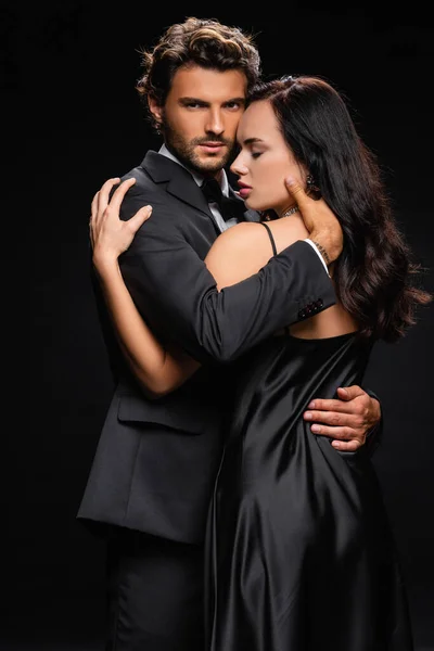 Leidenschaftlicher Mann blickt in die Kamera, während er elegante brünette Frau isoliert auf Schwarz umarmt — Stockfoto