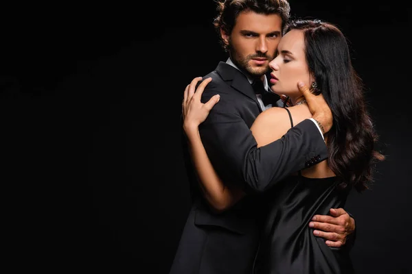 Apasionado hombre de traje abrazando sensual mujer y mirando a la cámara aislado en negro — Stock Photo