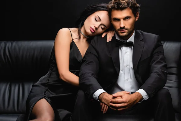 Mulher sexy em vestido de cetim deitado no ombro do homem elegante sentado no sofá de couro isolado no preto — Fotografia de Stock