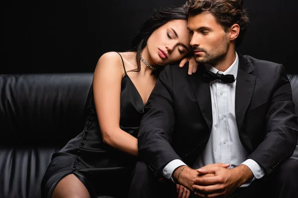 Elegant man sitting on leather couch near sensual woman leaning on his shoulder isolated on black — Stock Photo