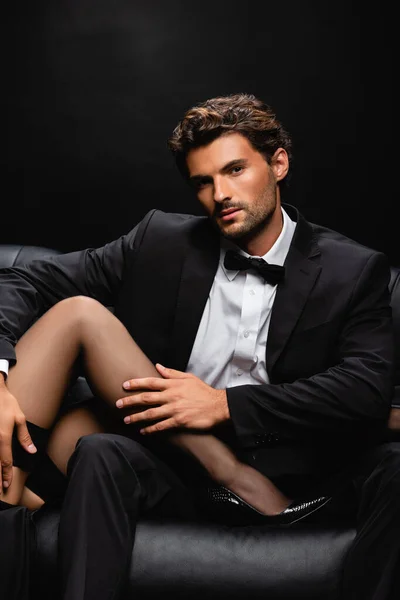 Young elegant man looking at camera while touching legs of seductive woman isolated on black — Stock Photo