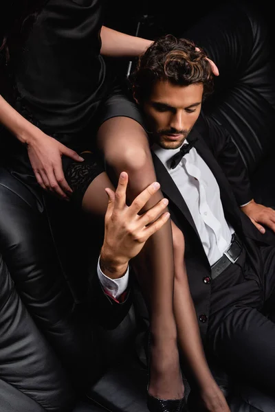 Элегантный мужчина в черном костюме касается ног сексуальной женщины, сидящей над ним на кожаном диване — стоковое фото