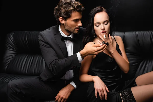 Hombre traje relámpago cigarrillo de mujer sensual en satén vestido negro aislado en negro - foto de stock