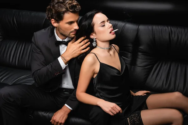 Man in suit touching hair of sexy woman smoking cigarette on leather couch on black — Stock Photo
