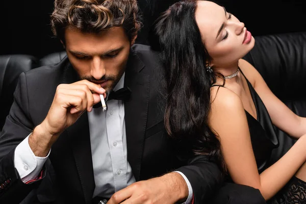 Seductive brunette woman with closed eyes leaning on elegant man smoking cigarette isolated on black — Stock Photo