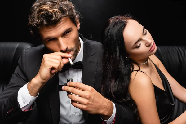 Hombre elegante cigarrillo relámpago y mirando a la cámara cerca de la mujer seductora sentado con los ojos cerrados aislados en negro - foto de stock