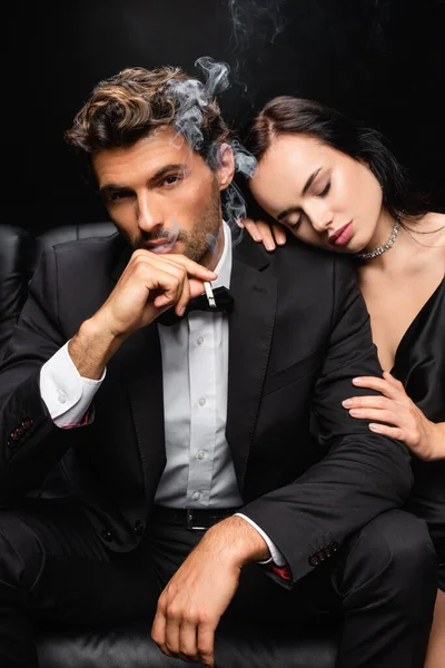 Mulher sensual com olhos fechados abraçando ombro de homem elegante fumando e olhando para a câmera isolada no preto — Fotografia de Stock