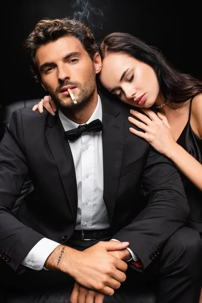 Seductora morena apoyada en el hombro del hombre fumando y mirando a la cámara aislada en negro - foto de stock