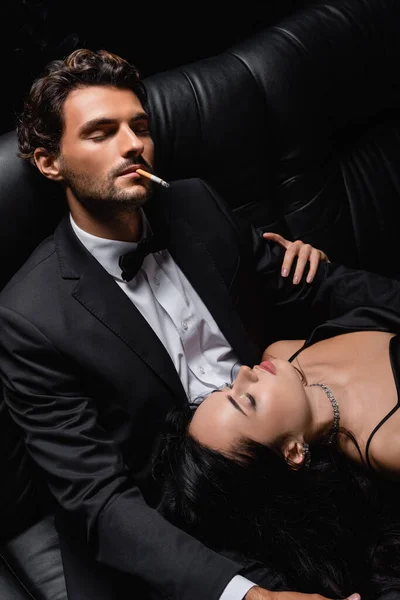 Man with closed eyes smoking cigarette near sexy woman lying on his laps — Stock Photo