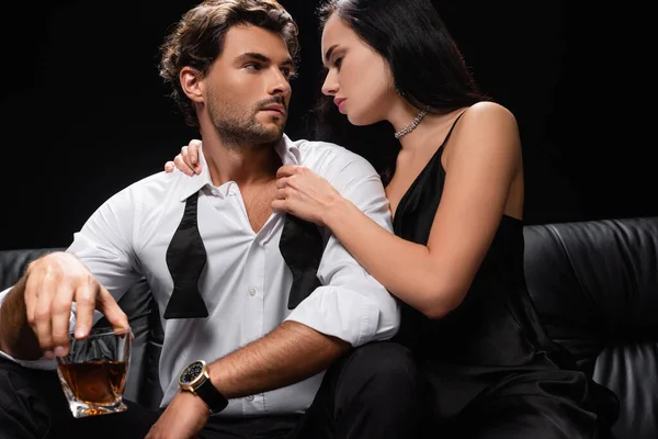 Sexy donna in abito di raso nero spogliarsi uomo in possesso di un bicchiere di whisky isolato su nero — Foto stock