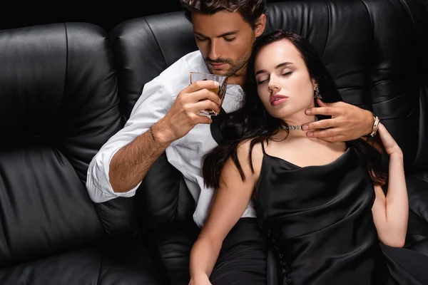 Man drinking whiskey and hugging neck of seductive woman in satin dress on leather couch isolated on black — Stock Photo