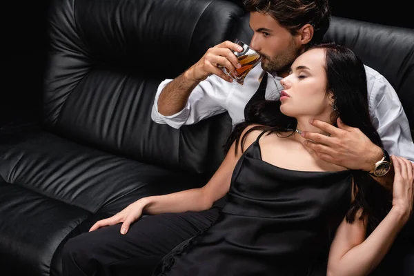 Jeune homme buvant du whisky tout en touchant le cou de la femme brune en robe satinée sur canapé en cuir noir — Photo de stock