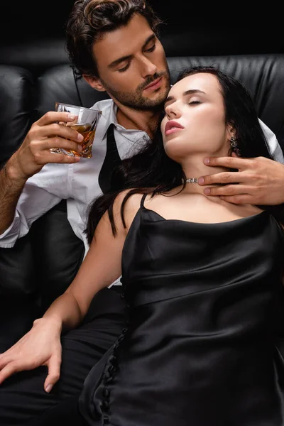 Young man holding glass of whiskey and embracing neck of seductive woman on black — Stock Photo