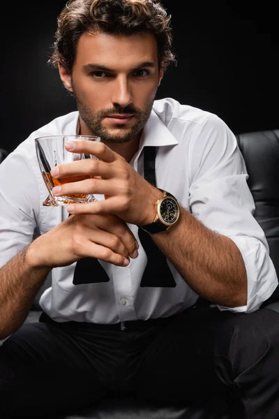 Hombre de camisa blanca y corbata desatada sosteniendo vaso de whisky y mirando a la cámara aislada en negro - foto de stock