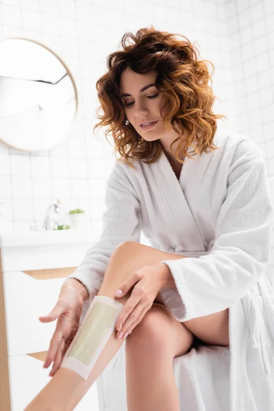 Woman with curly hair in bathrobe applying depilation stripe on blurred foreground in bathroom — Stock Photo