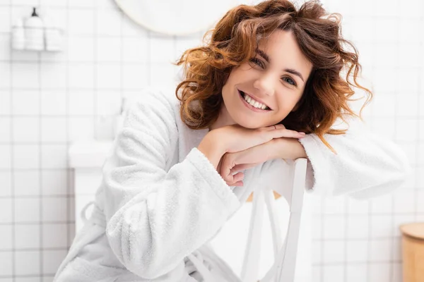 Femme gaie en peignoir regardant la caméra sur la chaise dans la salle de bain — Photo de stock