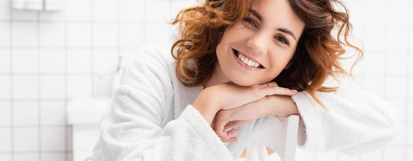 Кучерява жінка посміхається на камеру на стільці у білій ванній кімнаті, банер — стокове фото