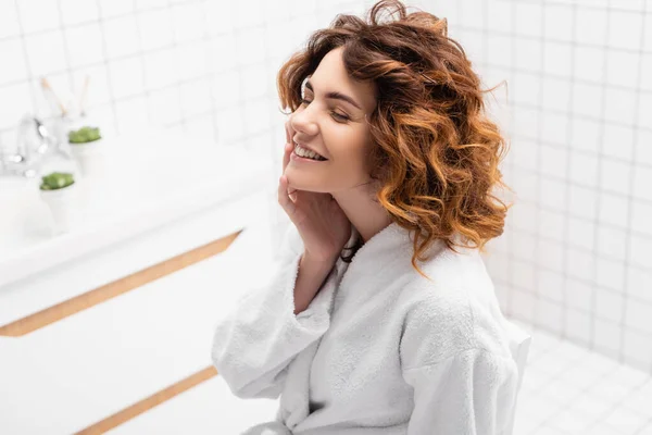 Femme en peignoir avec la main près du visage assis avec les yeux fermés dans la salle de bain — Photo de stock