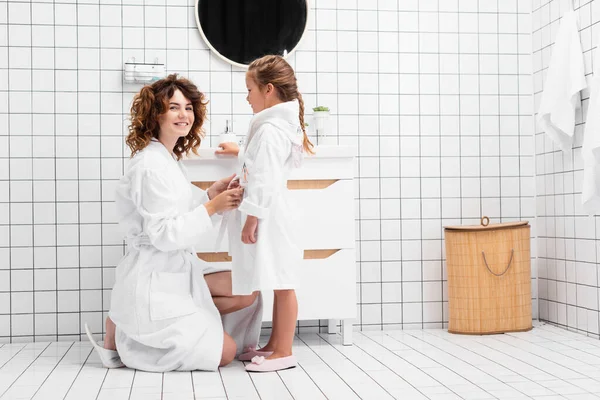 Улыбающаяся женщина в халате на дочери в ванной комнате — стоковое фото