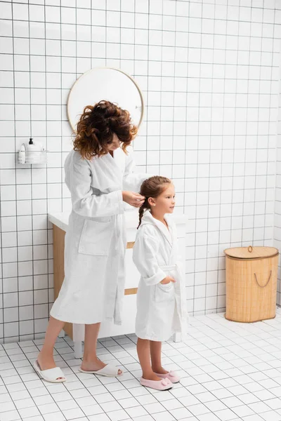 Madre en albornoz tejiendo el pelo del niño en el baño - foto de stock