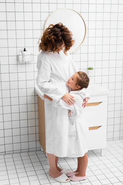 Ragazzo sorridente in accappatoio che abbraccia e guarda la madre in bagno — Foto stock