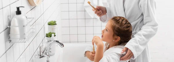 Niño en albornoz sosteniendo el cepillo de dientes cerca de la madre y fregadero en el baño, pancarta - foto de stock