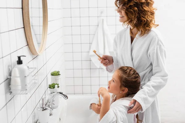 Улыбающаяся женщина с зубной щеткой рядом с ребенком и раковиной в ванной комнате — стоковое фото