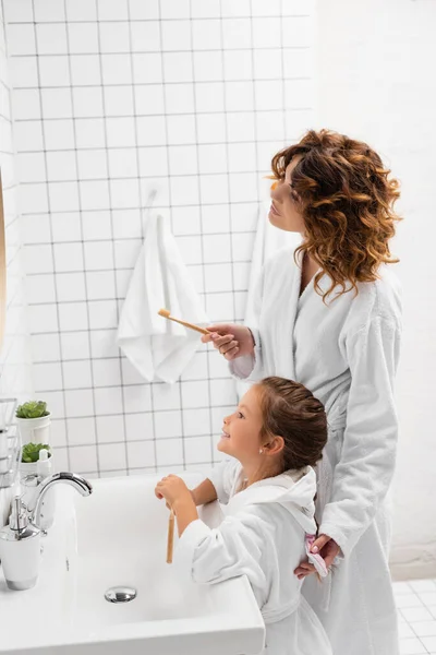 Madre e hija en albornoces sosteniendo cepillos de dientes cerca del lavabo - foto de stock