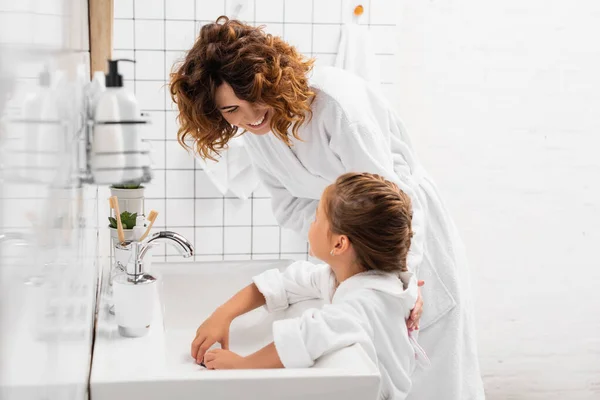 Улыбающаяся женщина обнимает дочь возле раковины в современной ванной — стоковое фото