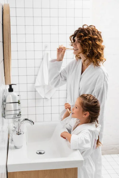 Mother and daughter in bathrobes brushing teeth near mirror — Stock Photo