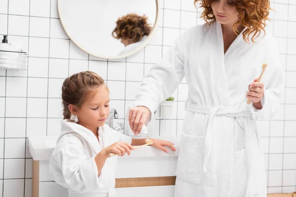 Hija sosteniendo cepillo de dientes cerca de la madre con pasta de dientes en el baño - foto de stock