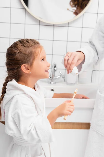 Sonriente niño sosteniendo el cepillo de dientes cerca de la madre con pasta de dientes en el baño - foto de stock