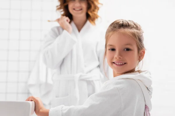 Smiling girl in bathrobe looking at camera near sink and mother on blurred background in bathroom — Stock Photo