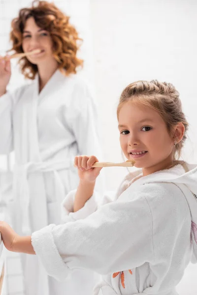 Smiling child in bathrobe brushing teeth near mother on blurred background — Stock Photo