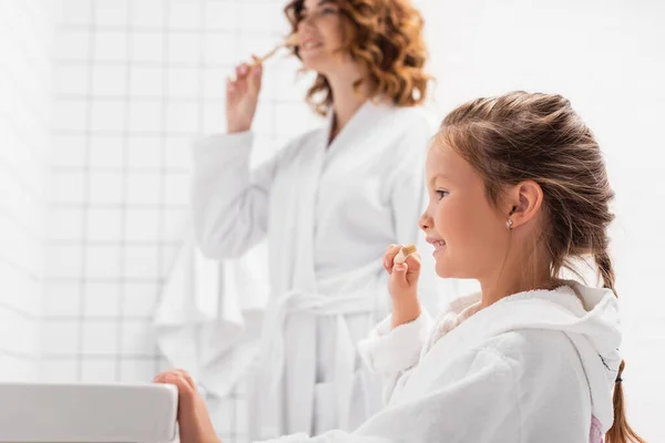 Cheerful girl in bathrobe brushing teeth near sink and mother on blurred background — Stock Photo