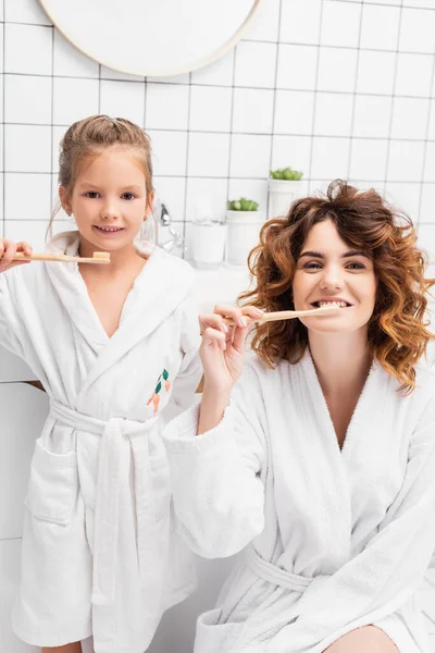 Mother and smiling child in bathrobes brushing teeth and looking at camera — Stock Photo