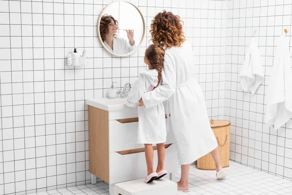 Mujer sonriente cepillándose los dientes y abrazando a su hija cerca del lavabo en el baño - foto de stock